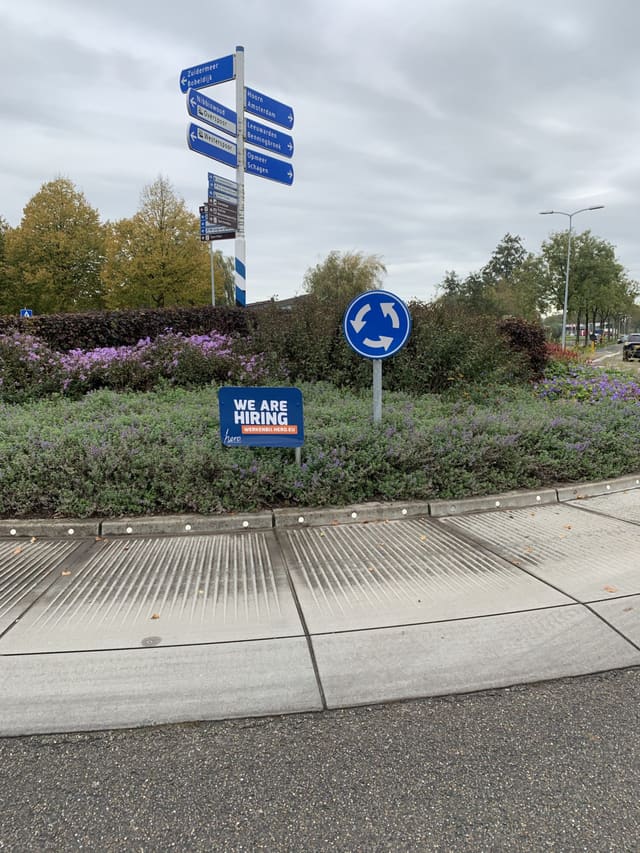 Rotonde met wegwijzers en een blauw vacaturebord tussen de beplanting