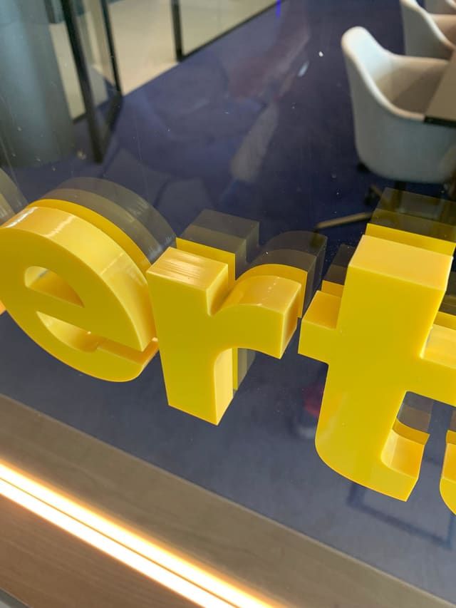 Gele 3D-letters van acryl op een glazen wand, met een kantoorruimte erachter