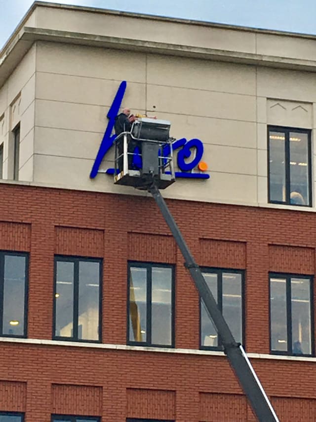 Blauwe 3D-letters worden vanaf een hoogwerker op de lichte bovenrand van een bakstenen pand gemonteerd