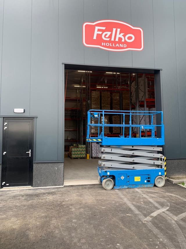 Rood logobord boven de deur van een bedrijfshal, met een schaarlift ervoor
