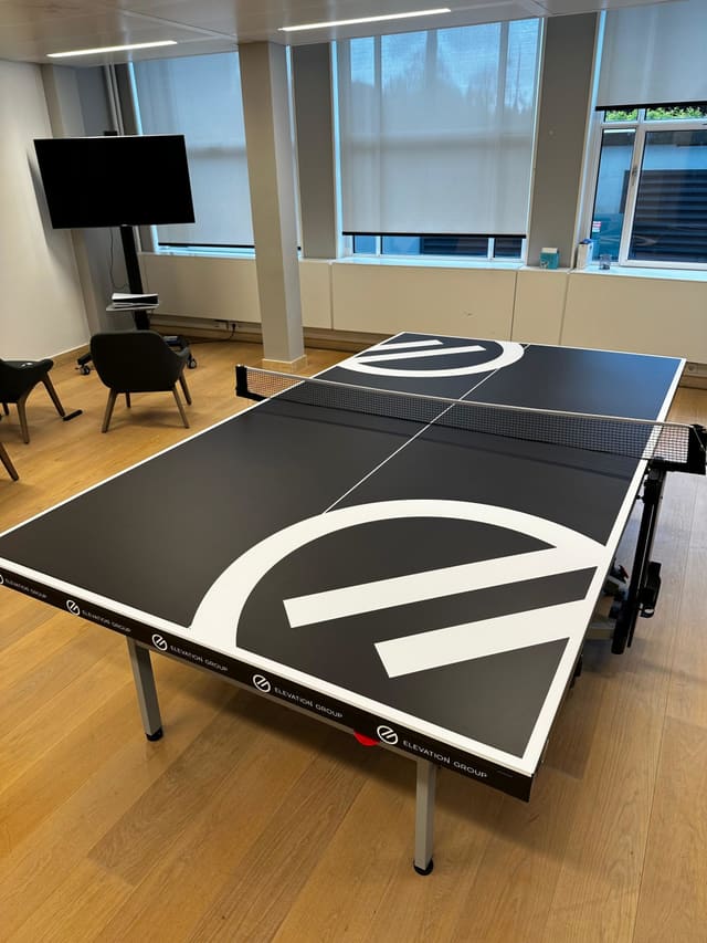 Zwarte pingpongtafel met een groot wit logo op het blad, in een kantoorruimte
