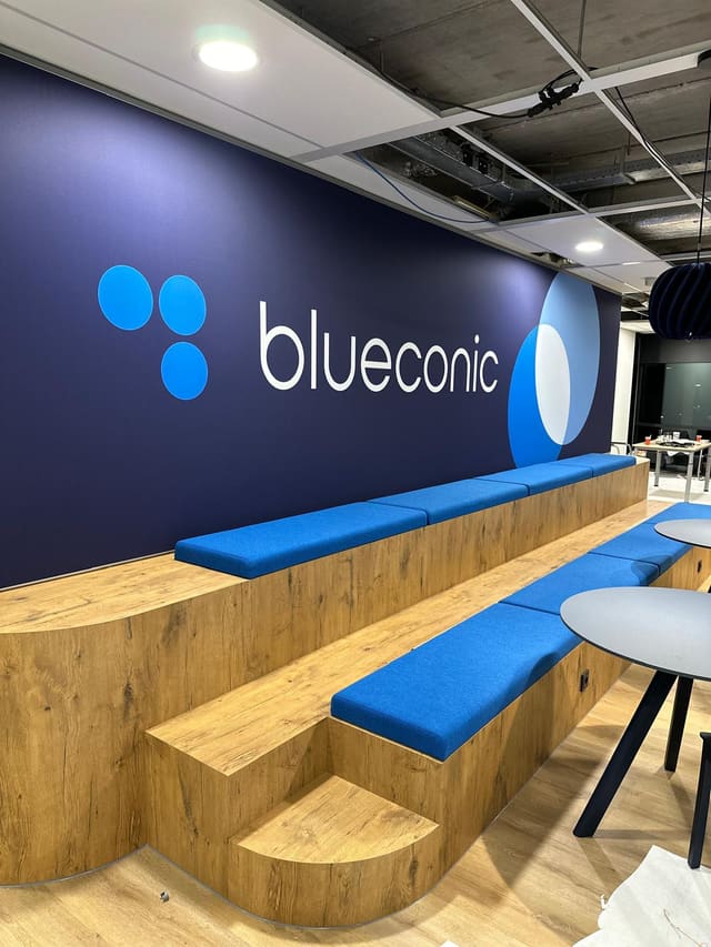 Donkerblauwe wand met een groot logo boven een houten zittribune met blauwe kussens