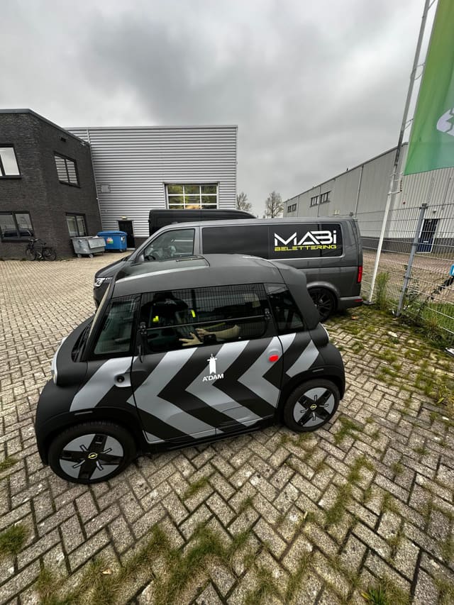 Kleine elektrische auto met een zwart-grijze chevronwrap; erachter staat de Mabi-bus